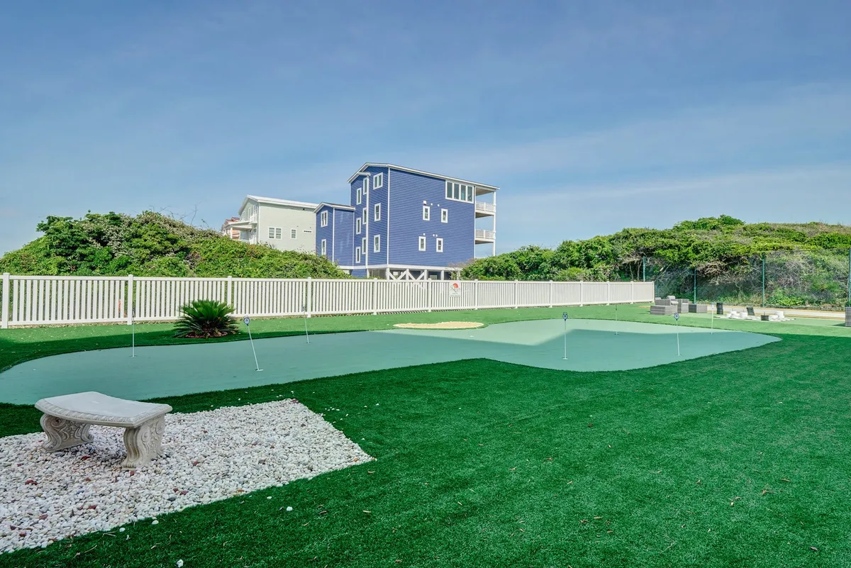 A lush green putting green with a white stone pathway and a blue house in the background, surrounded by greenery—offering a playful spot for friendly golf competitions or relaxed afternoons outside.