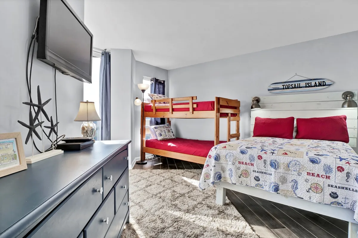 A playful and relaxed second bedroom showcasing a bed with red accent pillows, a twin-over-twin bunk bed, a television mounted on the wall, and a lamp on the nightstand. The room is designed for both families and groups, balancing comfort and fun.