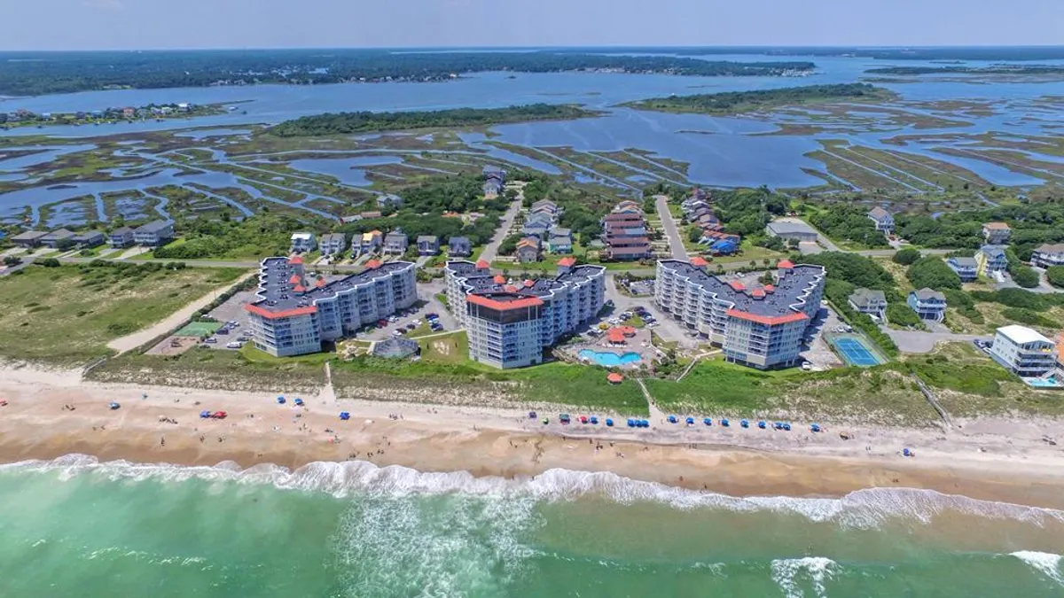 A vibrant beachfront scene with resort buildings, a large outdoor swimming pool, beach umbrellas, and the ocean beyond. The area is bustling with opportunities for swimming, sunbathing, and socializing, capturing the essence of a resort-style experience.