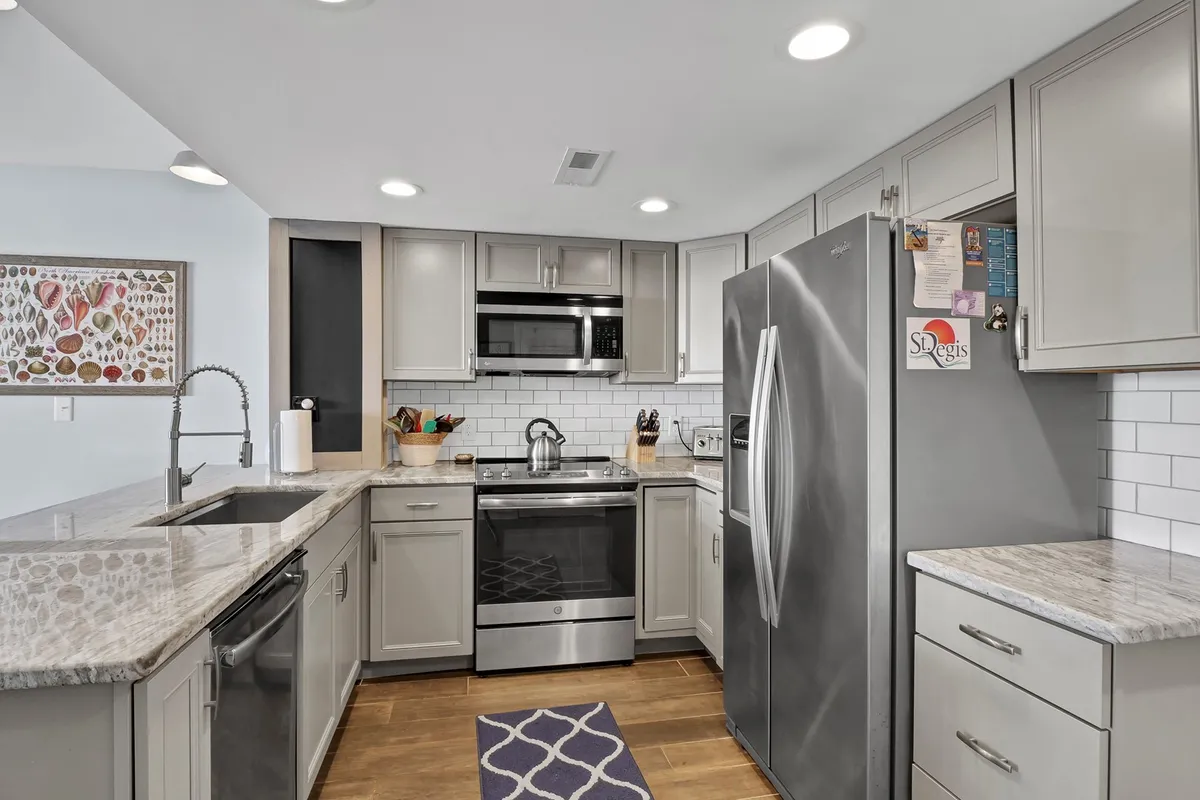 A warm and inviting kitchen featuring granite countertops, sleek stainless steel appliances, a modern tile backsplash, colorful wall art, and a cozy area rug. The space is clearly designed for both cooking and entertaining, with ample room for meal prep and family gatherings.