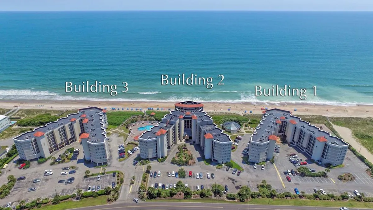 This exterior shot captures three condo buildings with a large swimming pool in the foreground, framed by vibrant blue ocean views in the distance. The scene embodies a quintessential beachside vacation atmosphere, where guests can enjoy both poolside relaxation and immediate access to the shoreline for romantic strolls.