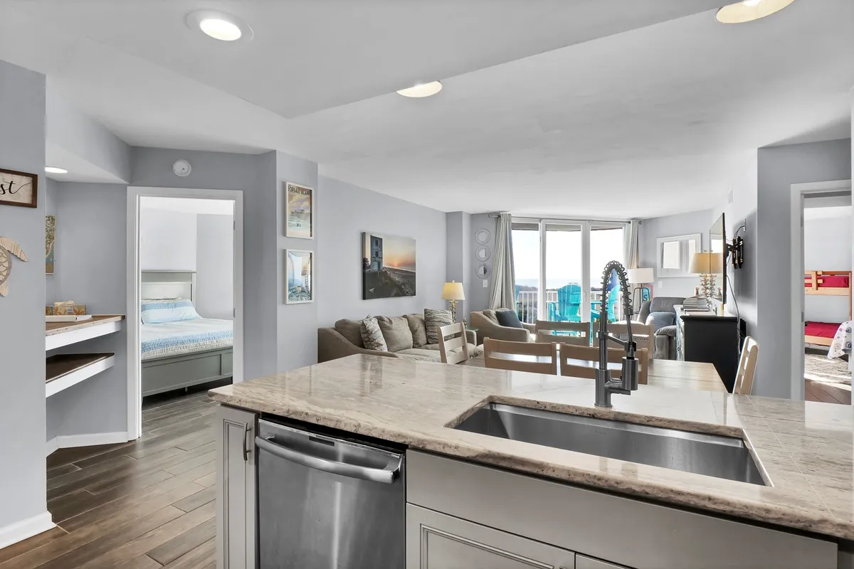 An open-plan living space that includes a kitchen island with sink and a welcoming lounge area with a sofa. Large windows reveal a scenic view, and stylish yet casual décor adds to the inviting, relaxed atmosphere. The design encourages gathering for meals, entertaining, and enjoying the vistas beyond the glass.