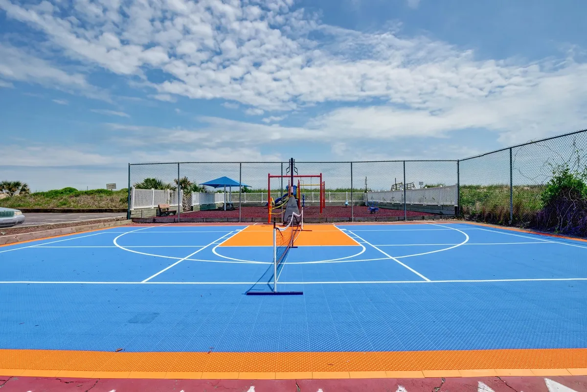 An outdoor area featuring a colorful basketball and tennis court with playground equipment visible in the background beneath a clear sky. The setting is vibrant and designed for both active recreation and play.
