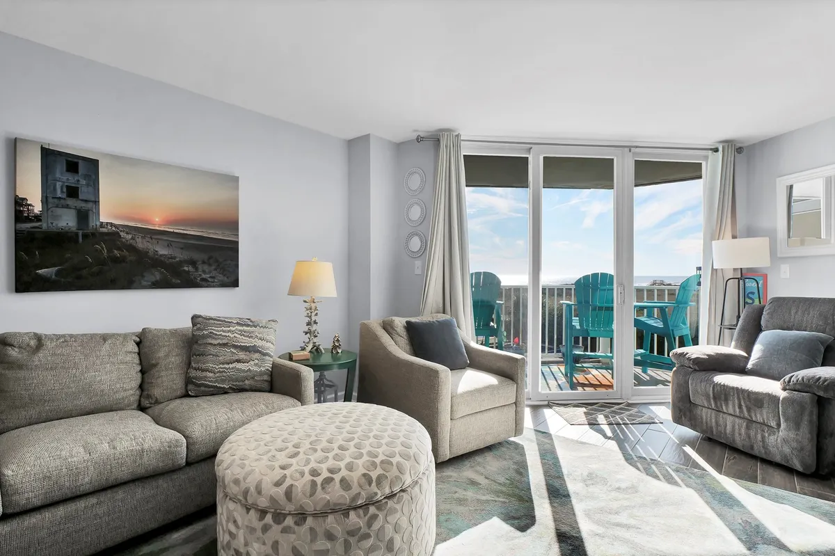 A cozy living area featuring a comfortable seating arrangement and large windows offering stunning ocean views. The space is filled with natural light, and soft beach-themed décor accents the room. Artwork on the wall and plush cushions invite relaxation while framing the striking seascape outside.