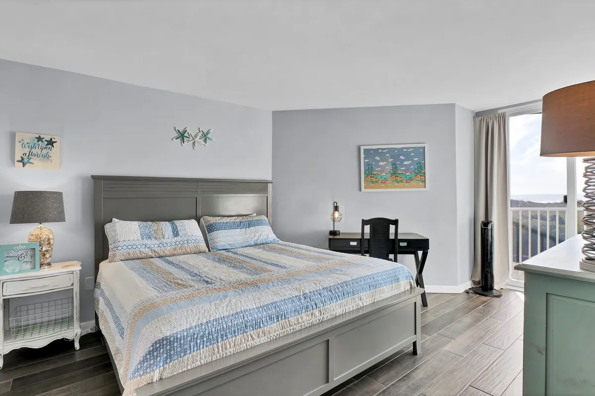 A calm and serene bedroom featuring a bed with a patterned quilt, a decorative lamp, artwork on the wall, and a desk with a chair. Sunlight softly fills the room, creating a tranquil atmosphere perfect for relaxation or enjoying a quiet morning coffee.