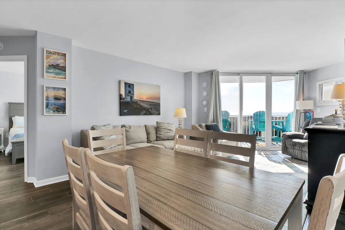 A bright living and dining space with a comfortable sofa, a dining table, and large windows that open to an expansive balcony. Sunlight pours into the room, illuminating the beach-inspired touches and inviting guests to relax or dine with a view.