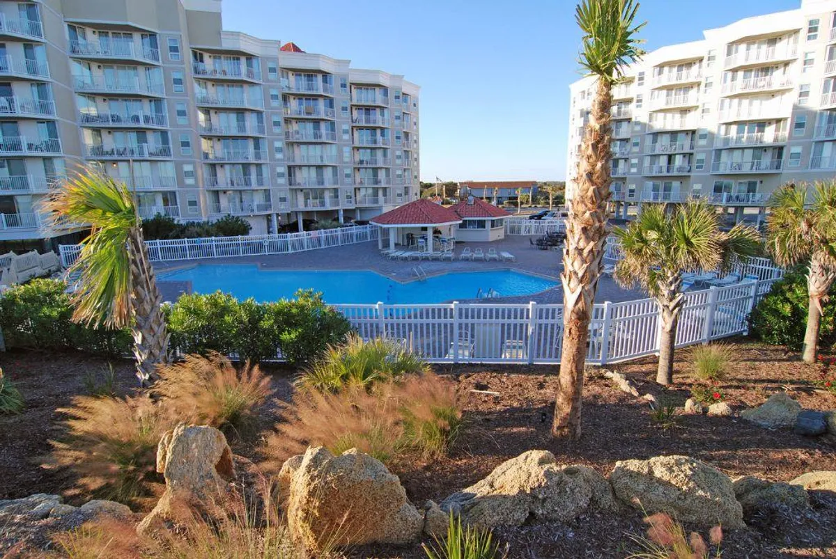 A sparkling swimming pool surrounded by palm trees and a rocky landscape, with the building facade in the background. The area is inviting for both sunbathing and social gatherings, evoking a leisurely resort atmosphere.