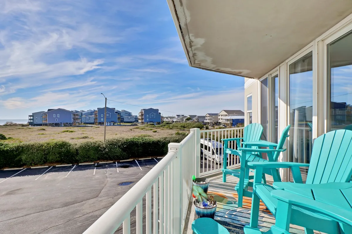 A pair of inviting turquoise chairs are positioned on a deck, offering a serene view of nearby beach houses and lush greenery. The chairs face outward, inviting couples to sit together and soak in the peaceful seaside ambiance.