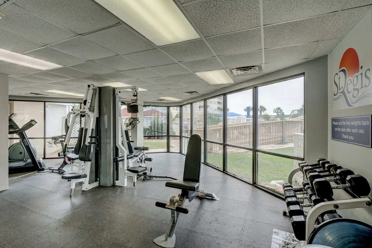A bright, modern fitness center features large windows that let in natural light and offer views outside. A variety of exercise machines and a weight rack are visible, creating a motivational setting for wellness and activity.