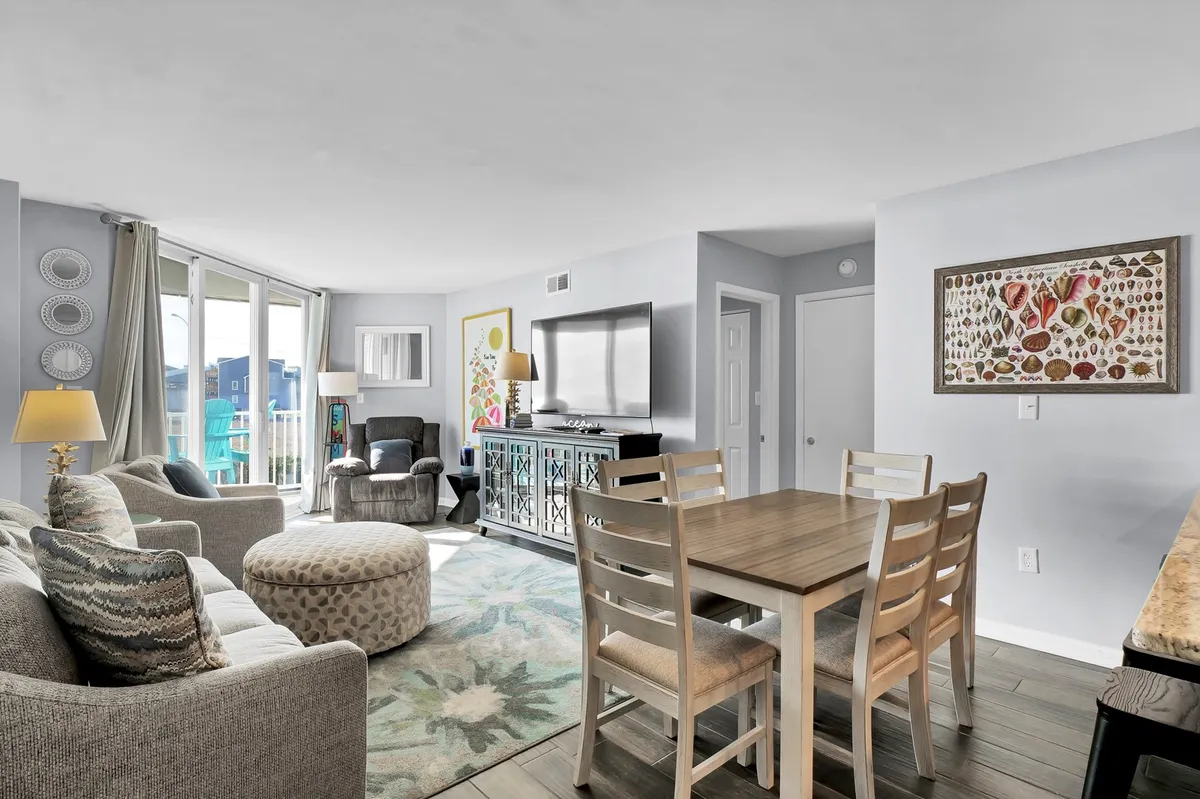 An inviting dining area with a spacious table set for meals, comfortable sofa seating close by, wall artwork, and large windows that let in natural light. The table is positioned to encourage group gatherings and lively meals, while the open layout connects the kitchen and living spaces.