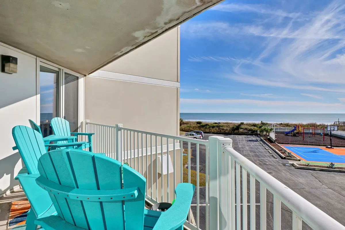 A tranquil outdoor area featuring turquoise Adirondack chairs with sweeping ocean views and a nearby playground area visible in the background. The setting is designed for both relaxation and play, with adults able to enjoy the view while children have fun close by.